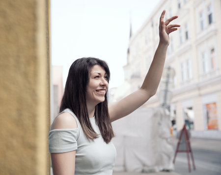 A smiling young woman waving her hand while standing in a bright and charming urban environmentの写真素材