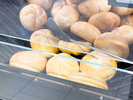 Variety of fresh bread rolls in bakery display with metal tongs.