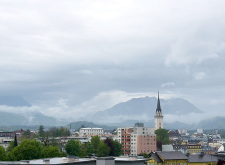 Cloudy urban landscape with church spire and mountainous background.の写真素材