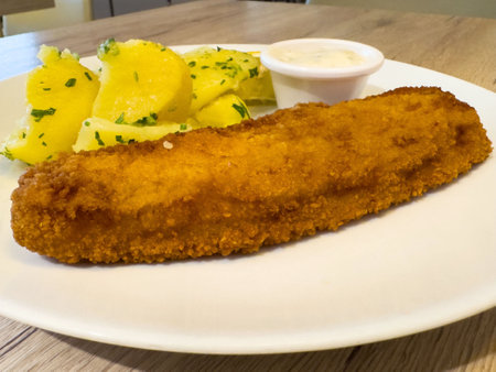 Crispy breaded fish fillet with herbed potatoes and dipping sauce on white plate.の写真素材
