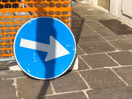 Blue right arrow traffic sign on sidewalk with orange barrier shadow in urban setting.