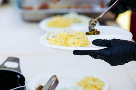 Close-up of hands serving creamy macaroni salad from plate at outdoor event.