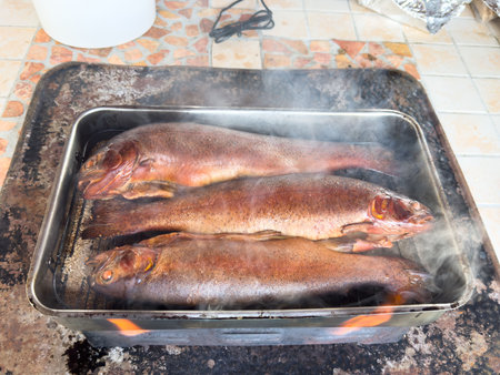 Whole trout are sizzling in a metal tray, releasing steam in a rustic kitchen, highlighting the art of cooking and the freshness of ingredientsの写真素材