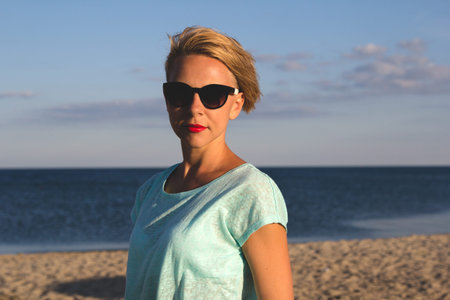 Outdoor photo of sexy beautiful elegant woman with short blonde hair resting on the summer beach next to the blue seaの写真素材