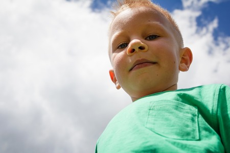 Cute little boy against blue sky with white clouds smiling and winking with cheeky look on his friendly faceの写真素材