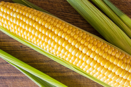 Ripe yellow corn cob with green leaves on brown wooden board