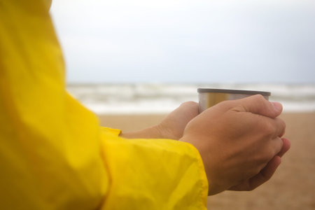 Men in yellow raincoat on the beach over the stormy sea holding a cup of hot drinkの写真素材