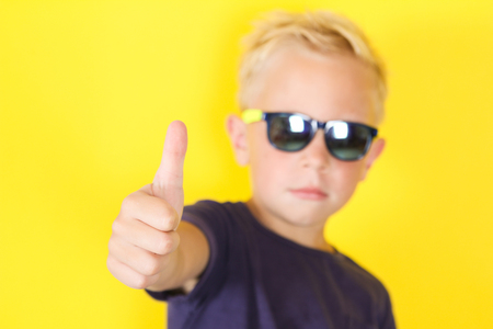 Cute blond boy wearing sunglasses on yellow background showing Thumbs Upの写真素材
