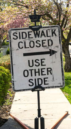 A sign notifies people of a closed sidewalk, with trees blooming with flowers in the backgroundの写真素材