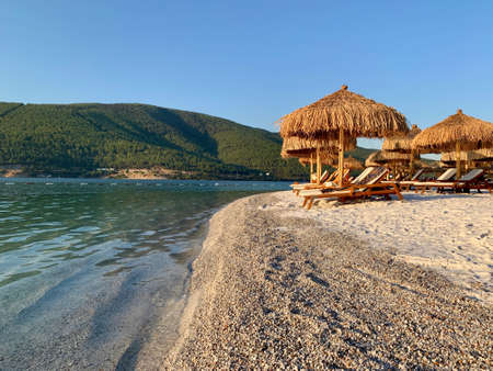 Chic view of the emerald Aegean coast with white sand, bamboo palms and sun loungers. Luxury leisure relaxation concept. White beach with a mountain in the backgroundの写真素材