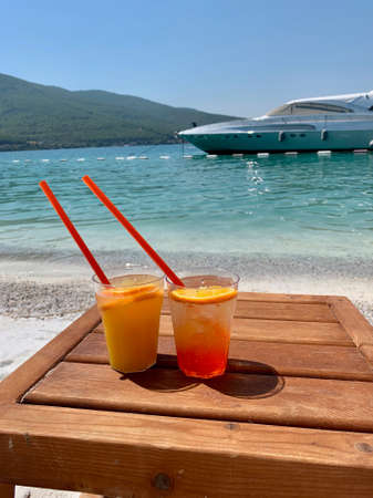 Two glasses of aperol with slices of lemon on a wooden tray against the background of the emerald Aegean Sea and a snow-white yacht. High quality photoの写真素材