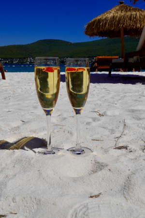 Two cold glasses of champagne with raspberry on white sand on the beach in an emerald bay. Luxury resort with sea view. Romantic vacationの写真素材