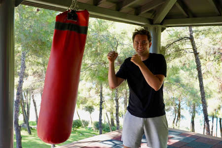 Joyful young man boxing a red pear against the background of a green forest on an outdoor in the street. Sports and health concept. Outdoor training. A young man is boxing in the gym.の写真素材