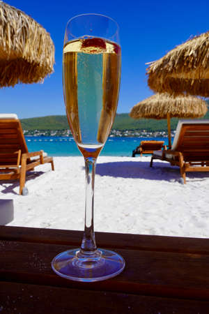 High glass of champagne on a thin leg with raspberry on white sand on the beach in an emerald bay. Luxury resort with sea view. Romantic vacationの写真素材