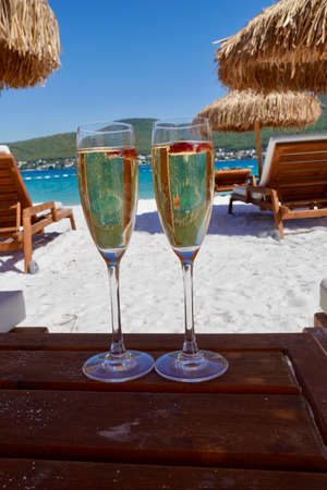 Two cold glasses of champagne with raspberry on white sand on the beach in an emerald bay. Luxury resort with sea view. Romantic vacationの写真素材