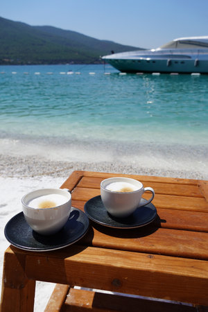 coffee white with grey saucer cup of cappuccino on a wooden table on the sea beach with an emerald view of the ocean, white boats backgroundの写真素材