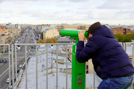 A young man in blue puffer looking through binoculars to the urban landscape from the roof top of Saint-Petersburgの写真素材