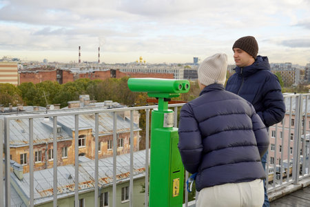 A young man in blue puffer looking through binoculars to the urban landscape from the roof top of Saint-Petersburgの写真素材