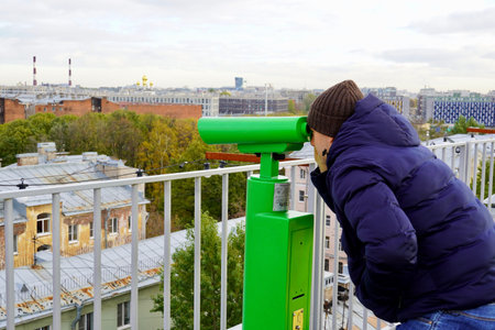 A young man in blue puffer looking through binoculars to the urban landscape from the roof top of Saint-Petersburgの写真素材