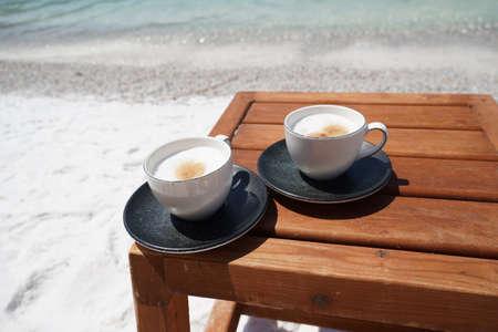 White cup of coffee cappuccino with grey saucer on a wooden table on the sea beach with an emerald view of the ocean, white boats in background, wonderful see viewの写真素材