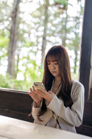 Scene of a female college student in a white jacket sitting on a chair in the forest and operating a smartphone.の写真素材