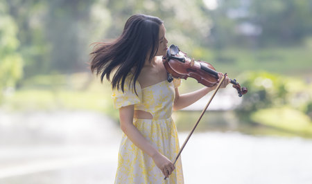Twenty-something Chinese woman in yellow dress gracefully playing violin in the forest of a large, nature-filled park in Bukit Jalil, near Kuala Lumpur, Malaysia.の写真素材