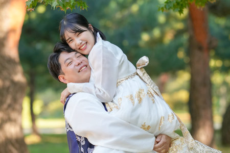 A Korean couple, a man in his 30s and a woman in her 20s, are wearing Hanbok and hugging each other in a park surrounded by green trees in Seoul, South Korea.の写真素材