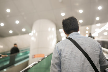 Man wearing a blue shirt and a black backpack is walking down an escalator. There are other people in the backgroundの写真素材