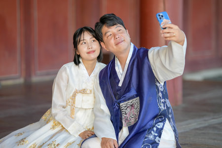 A Korean couple, a man in his 30s and a woman in her 20s, are happily wearing Hanbok and taking a selfie with a smartphone inside a historical building in Seoul, South Korea.の写真素材