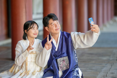 A Korean couple, a man in his 30s and a woman in her 20s, are happily wearing Hanbok and taking a selfie with a smartphone inside a historical building in Seoul, South Korea.の写真素材