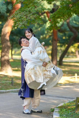 A Korean couple, a man in his 30s and a woman in her 20s, are wearing Hanbok and hugging each other in a park surrounded by green trees in Seoul, South Korea.の写真素材