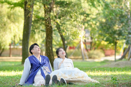 A Korean couple in their 30s and a woman in their 20s are looking at the sky in a green park surrounded by trees while wearing Hanbok in Seoul, South Korea.の写真素材