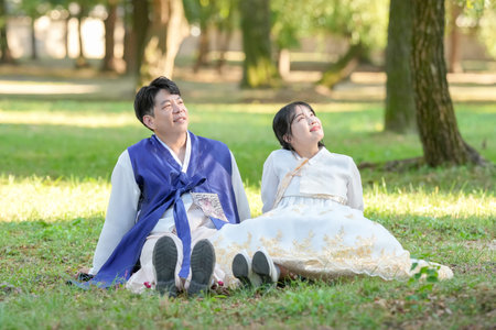 A Korean couple in their 30s and a woman in their 20s are looking at the sky in a green park surrounded by trees while wearing Hanbok in Seoul, South Korea.の写真素材