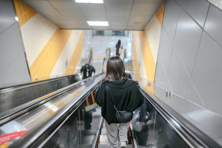 Woman is walking down an escalator. She is wearing a black hoodie and carrying a black handbagのeditorial素材