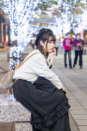 A young and cute Taiwanese woman in her 20s wearing black and white clothes, sitting and resting at night amidst beautiful illuminations in a shopping mall in Xinyi District, Taipei, Taiwanの写真素材