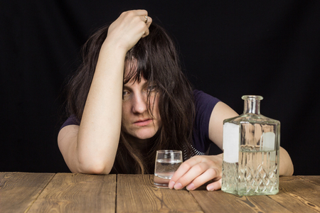 Drunk girl with a shot of alcohol and a bottle of alcohol, black background, wooden tableの写真素材