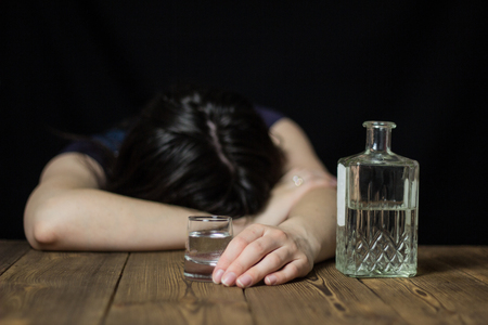 A drunk girl laid her head on a table, in her hand a glass of alcohol and a bottle, a wooden table, a black background, female alcoholismの写真素材