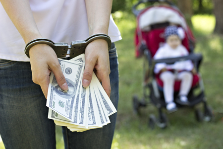 A girl is holding money dollars in handcuffs against the backdrop of a baby in a carriage, arrest, detention for sale by children, trafficking in children, close up, little babyの写真素材