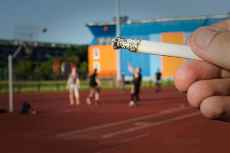 A man holds a cigarette in the background of the stadium where they play basketball, close-up, bad habit, healthy lifestyleの写真素材