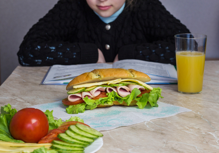 The girl does the lessons before her on the table lies a sandwich, vegetables and juice, caucasianの写真素材