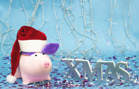 Soft toy pink pig in a red Santa Claus hat on the background of the Christmas tree on a blue background, the inscription xmas, the explosion of the Christmas crackers and Christmas tinsel and candy, copy space, concept, 2019の写真素材