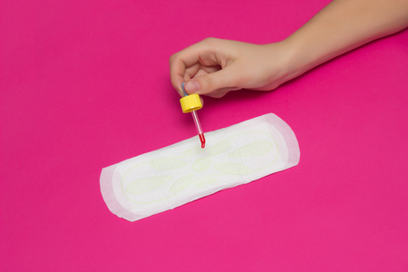 A female hand holds a pipette in her hand and drips blood onto a sanitary pad, menstruation concept, pink background, copy space, protectionの写真素材