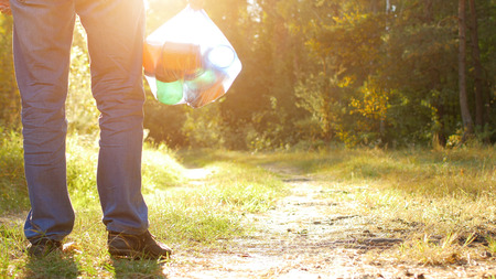 A man with a package of garbage in his hands with plastic in the forest against the backdrop of a forest road, a sunset, copy space, trashの写真素材