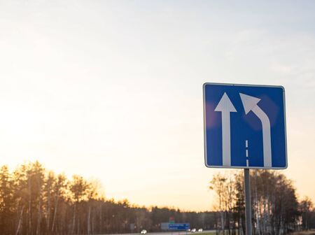 Blue road sign on sky background with sunny sunset narrowing the road to the right. Concept of...