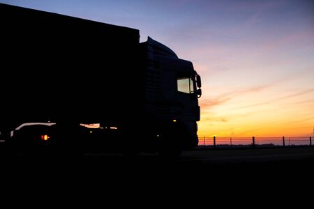 A truck with a trailer rides on the motorway at night against the backdrop of an orange sunny sunset. The concept of the regime of work and rest of truckers, copy spaceの写真素材