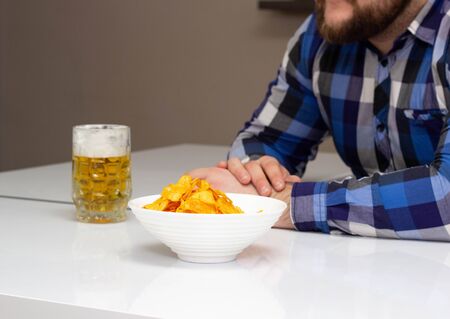 Bearded man drinks beer and eats chips in the kitchen, junk food, togethernessの写真素材