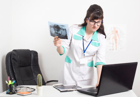 Doctor radiologist holds an x-ray picture of the tooth jaw and checks the data in a laptop, doctor's office in the clinic. Radiation Diagnostics with X-Rayの写真素材
