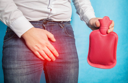 A man in a white shirt holds his groin and holds a red heating pad with hot water in his hand. The...