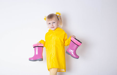 A little girl aged 3 years in a yellow raincoat holds pink rubber boots in her hands on a white background, isolate. The concept of bad weather and autumn.の写真素材