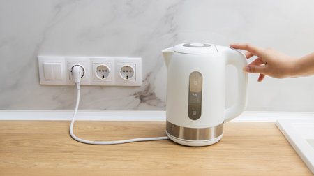 A woman turns on an electric kettle to boil water in the kitchen. Electricity consumption by household appliances, electricity consumption. Copy space for textの写真素材